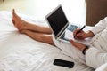 a man in a bathrobe with a laptop on his lap lies in the bed and writes something in a notebook.