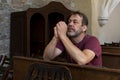 A man with a beard, 50-55 years old, prays in a church on a bench, his hands are folded in prayer, he looks straight.