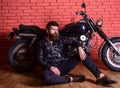 Man with beard, biker in leather jacket near motor bike in garage, brick wall background. Brutal biker concept. Hipster
