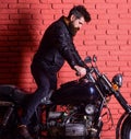 Man with beard, biker in leather jacket near motor bike in garage, brick wall background. Hipster, brutal biker on