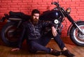 Man with beard, biker in leather jacket near motor bike in garage, brick wall background. Hipster, brutal biker on