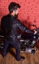 Man with beard, biker in leather jacket near motor bike in garage, brick wall background. Masculine hobby concept