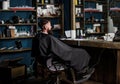 Man with beard covered with black cape sits in hairdressers chair in front of mirror. Man with beard client of hipster