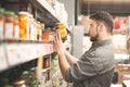 Man from a beard is in the department of canned vegetable supermarket, holds a bottle of tomatoes and reads a label. Buyer selects