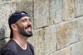 Man with beard leaning on a stone wall, smiling, happy, dreaming, looking up away