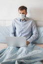 Young man with a beard in medical mask in a shirt sits in bed with a laptop and works from home