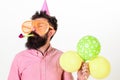 Man with beard and mustache on calm face holds air balloons, white background. Guy in party hat with party horn Royalty Free Stock Photo