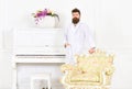 Man with beard and mustache enjoys morning while stand near piano and old fashioned armchair. Man sleepy in bathrobe