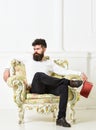 Man with beard and mustache sits on armchair, holds book, white wall background. Connoisseur on thoughtful face finished