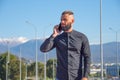 Man with a beard and slicked back hair in black shirt talking on phone, stands on the street of the city from behind against the