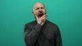 Man with a beard yawning against a solid green background, showcasing a bald head and grey sweater, exuding a handsome, tired Royalty Free Stock Photo