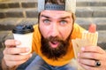 Man bearded enjoy quick lunch stairs background. Hipster eat hot dog. Hipster enjoy hot dog drink paper cup. Fast food