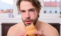 Man bearded handsome guy eating breakfast in bed. Guy holds croissant bedroom hotel room window background. Man eats