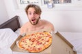 Man bearded handsome guy eating cheesy food for breakfast in bed. Guy holds pizza box sit bed in bedroom or hotel room