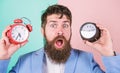 Man bearded hipster hold two different clocks. Guy unshaven puzzled face having problems with changing time. Time zone