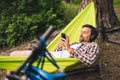 Man on bicycle trip at camping by lake is relaxing in green hammock while listening to music. Active recreation theme in Royalty Free Stock Photo