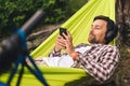 Man on bicycle trip at camping by lake is relaxing in green hammock while listening to music. Active recreation theme in Royalty Free Stock Photo