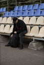 Man in black bomber jacket and cap sitting alone in empty stadium seating area with a backpack Royalty Free Stock Photo