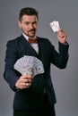 Man in black classic suit and red bow-tie showing two playing cards and cash, posing on gray studio background. Gambling Royalty Free Stock Photo