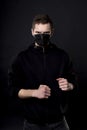 Man in black in a fighting pose with clenched fists. On the young man`s face, a black medical mask covers his nose and Royalty Free Stock Photo