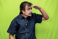 A man in black shirt against a green screen, shading his left hand above his eyes to see something on the left amidst harsh light