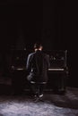 Man in black suit sit in front of piano in black interior, rear view