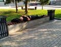 A man in a black t-shirt and pants lying on a wooden bench. Hands with a clock folded on his chest. Next to his head is a jacket.