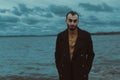 A man in a black trench coat stands on the shore of a cold lake.