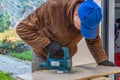 A man in the black working gloves and a brown jacket and a blue hat cuts a board using a Jigsaw power tool on a work