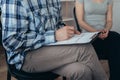 Man in a blue checked shirt and grey pants sits on a chair and holds a writing pad on his lap