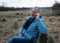 Man in a blue coat with the mobile phone sitting on the bench in a field