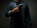 A man in a blue denim shirt is standing in a dark room holding a pistol to his chest. concept of assassination, murder, criminal
