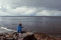 Man in blue jacket and a hat sitting on a rock by the ocean. Back to camera. Storm clouds over water surface. Dark depressive Royalty Free Stock Photo