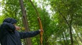 A man in a blue jacket, pulls the bowstring, to hit the target, against the green forest