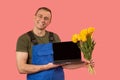Man in blue jumpsuit holds bouquet of tulips and laptop with blank black screen and smile. Concept of buying flowers online. Copy Royalty Free Stock Photo