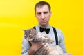 Man in blue shirt, black suspender and bow tie holding grey striped tabby cat with outstretched paws and looking at camera