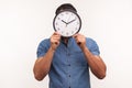 Man in blue shirt hiding face behind wall clock display, wasting his time, procrastination, organization of working time Royalty Free Stock Photo