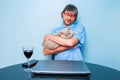 Man in blue shirt holding cute chubby British short hair cat in his hands sitting at a table with computer and glass of red wine. Royalty Free Stock Photo