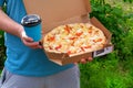 Man in blue t-shirt is holding box with tasty pizza and paper cup with drink, close up view. Italian Cuisine. Snack on a sunny Royalty Free Stock Photo