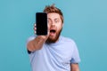 Man in blue T-shirt holding out cellphone and looking at camera with big eyes and open mouth. Royalty Free Stock Photo