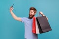 Man in blue T-shirt holding shopping bags and talking selfie on smartphone, satisfied with purchase. Royalty Free Stock Photo