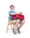 Man in a blue T-shirt, shorts and sunglasses sits on a chair with a red suitcase waiting. Isolated on white background Royalty Free Stock Photo