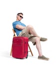 Man in a blue T-shirt and white shorts sits on a chair near a red suitcase, waiting. Isolated on white background Royalty Free Stock Photo