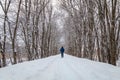 Man in blue walking alone on snow trail under tall tree canopy in winter