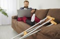 Man with broken leg using laptop computer while sitting on couch with crutches at home