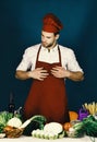 Man in burgundy cook hat and apron. Cook works in kitchen near table with vegetables and tools. Royalty Free Stock Photo