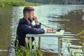 A man in a business suit is sitting at a white table in a swamp, is looking at a laptop and talking on the phone