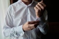 Man buttoning cuff link of luxury white shirt sleeve, copy space. Groom fixes cufflinks standing near window.