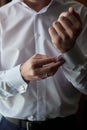 Man buttoning cuff link of luxury white shirt sleeve, copy space. Groom fixes cufflinks standing near window.