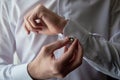 Man buttoning cuff link of luxury white shirt sleeve, copy space. Groom fixes cufflinks standing near window.